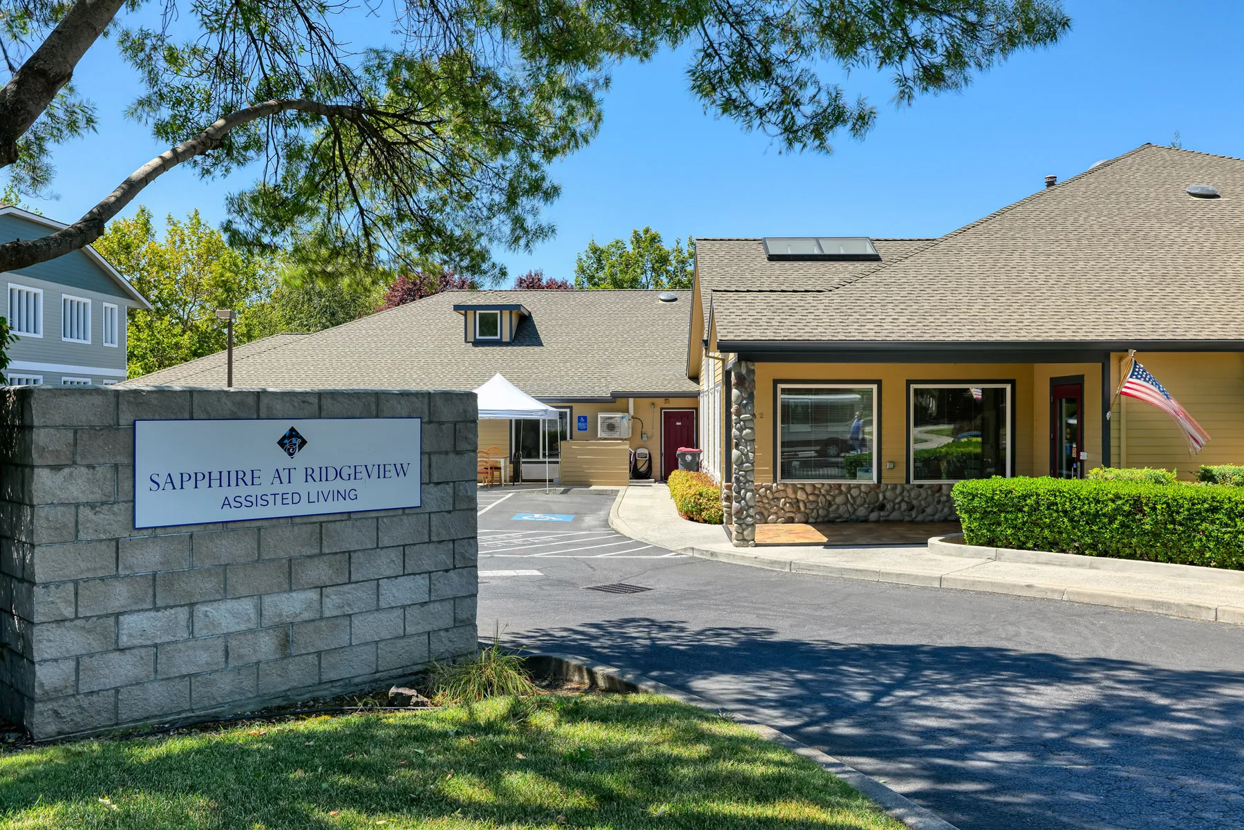 Exterior view of Sapphire at Ridgeview Assisted Living facility, featuring a sign, well-maintained landscaping, and an American flag Exterior view of Sapphire at Ridgeview Assisted Living facility, featuring a sign, well-maintained landscaping, and an American flag